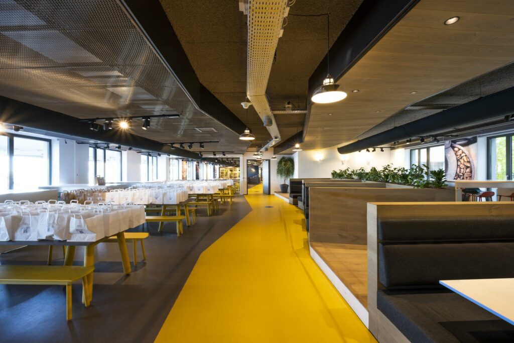 Moderne bedrijfskantine aan de Piet Heinkade met gele vloer, lange tafels vol lunchpakketten en zithoeken met planten.